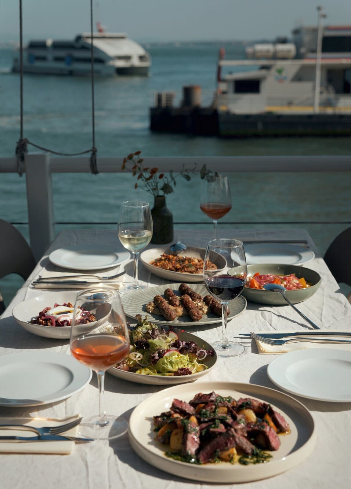 A table laid at TEJÁ on the terrace, dishes and glasses along it, the river and the ferries behind.
