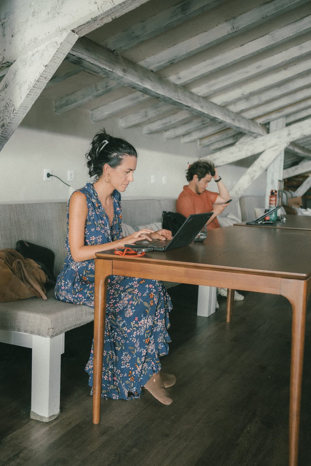 A woman in a blue floral dress typing on a laptop at a long wooden table, a man on the bench behind her.