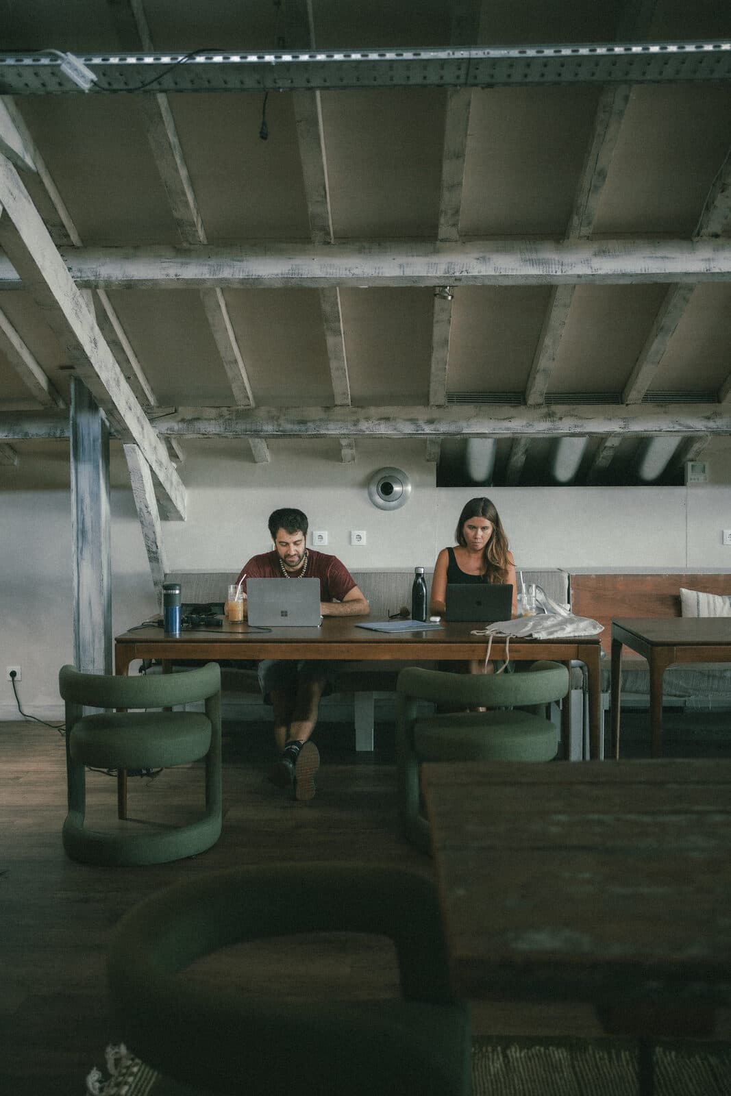 A man and a woman working side by side on laptops at a long wooden table under a beamed ceiling.