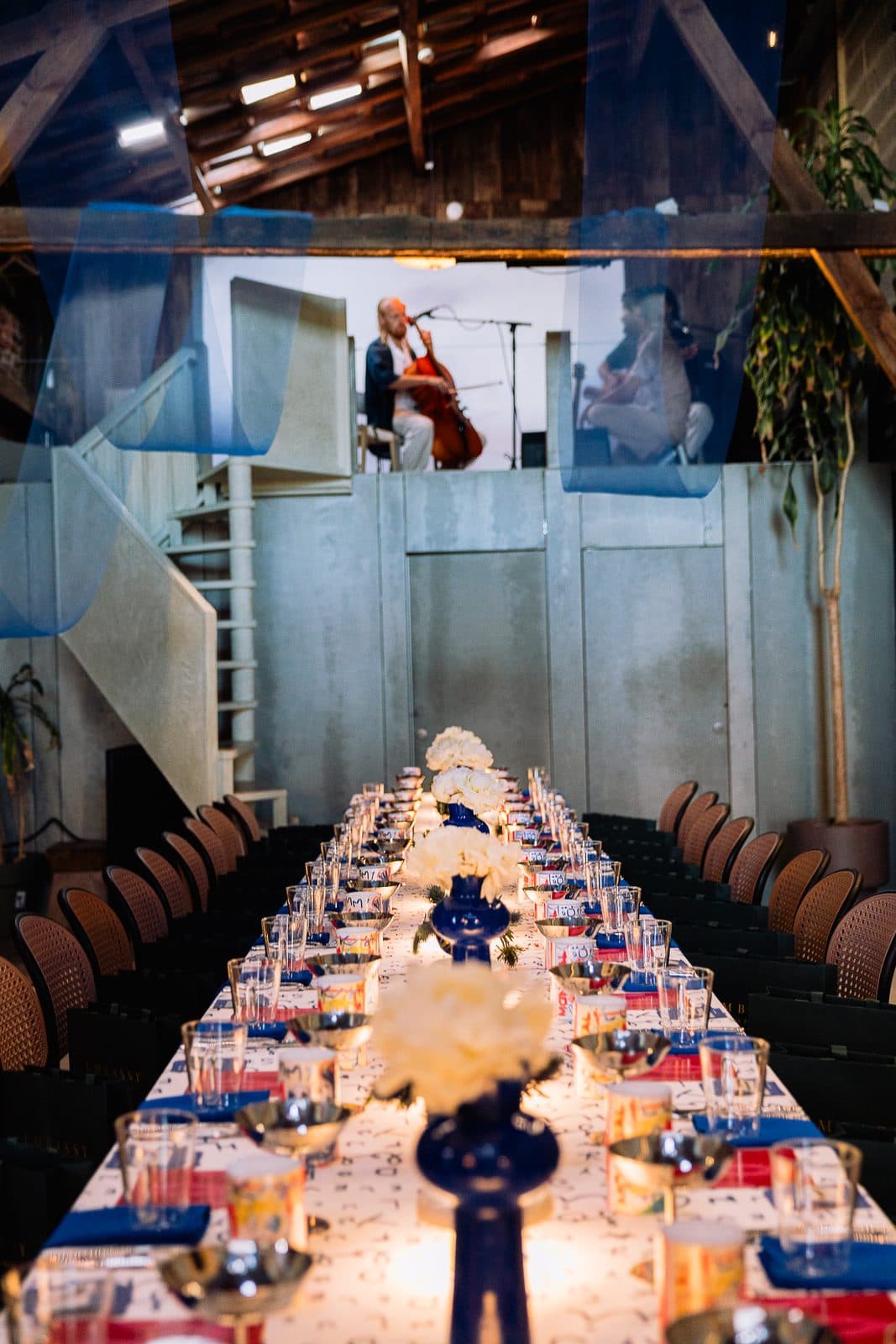 A laid long table running toward a raised platform at the end of the hall, where a cellist and a singer are playing.
